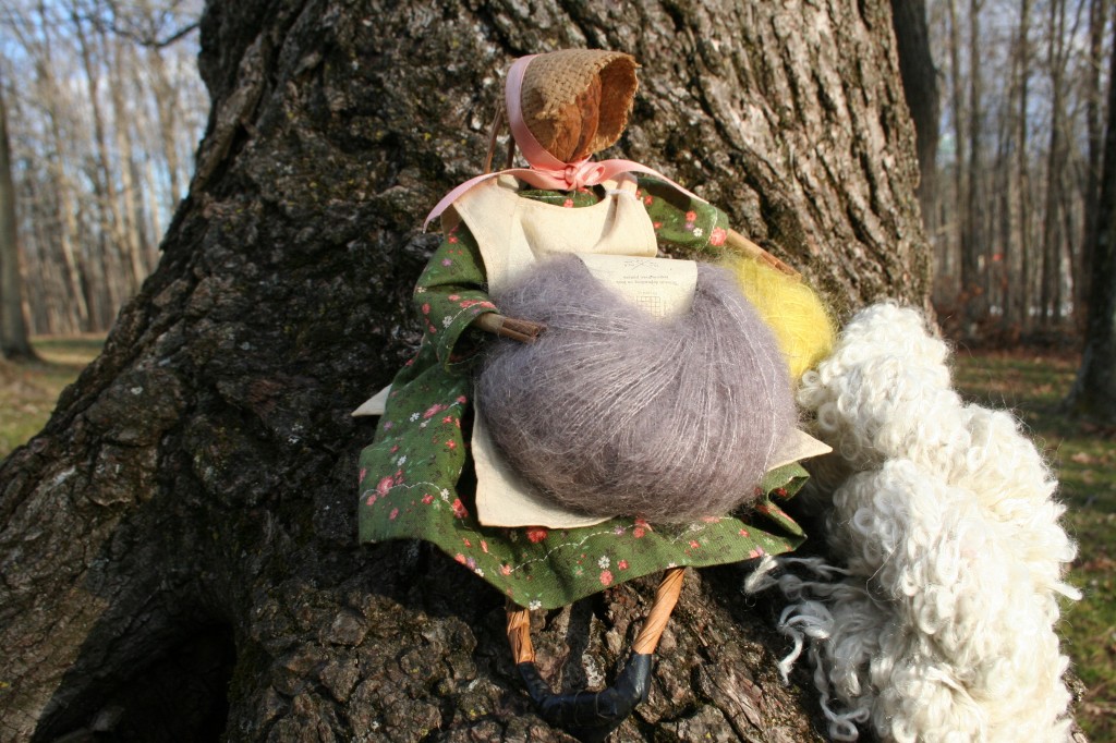 A handmade mountain folk figure with a walnut for a head sits with a cake of kid mohair blended with silk, a wound kid mohair/silk yarn ball in  a cream-ecru loopy mohair skein.